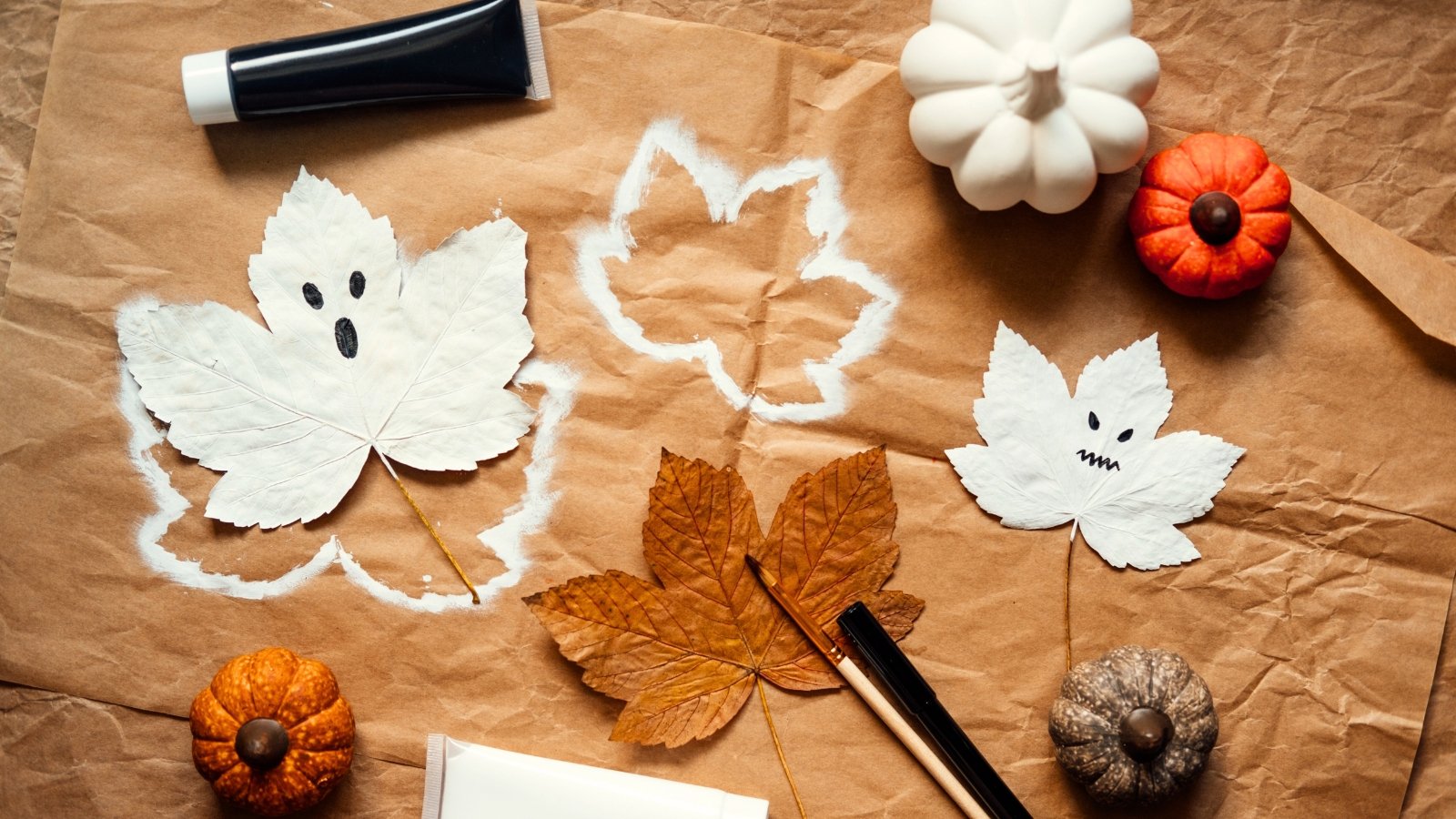 An artful arrangement of decorative white and orange pumpkins, scattered orange maple leaves, and several white-painted leaves shaped like Halloween ghosts.